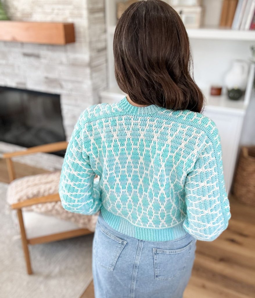 A woman with long brown hair stands indoors, facing away, wearing a Seaglass mosaic cardigan sweater and high-waisted blue jeans.