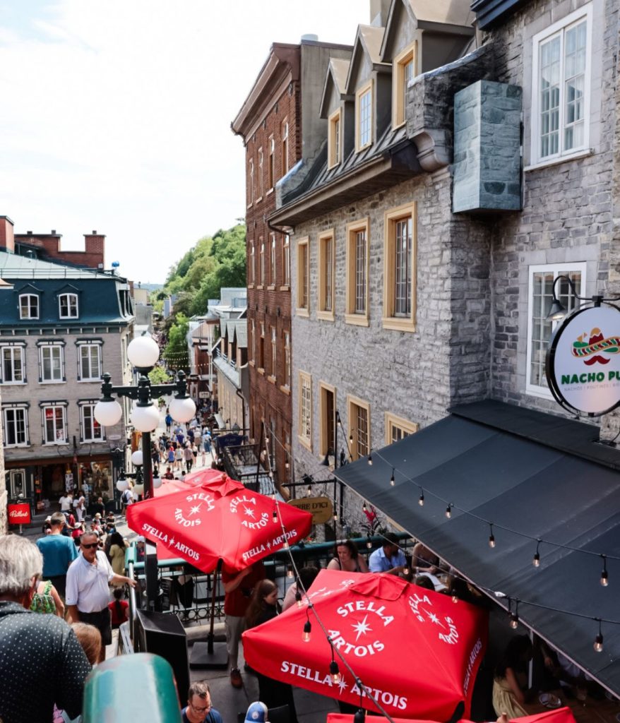 A busy street scene with people walking, outdoor seating under red umbrellas, and someone wearing a stylish Granny Stitch Top amid historic stone buildings in a downtown area.