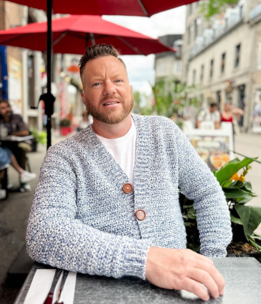 Man with a trimmed beard wearing a blue and white men's crochet cardigan sits at an outdoor café table under a red umbrella on a city street.