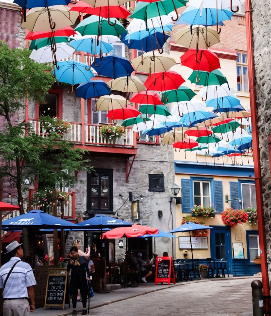 Outdoor café scene in a narrow street with colorful umbrellas suspended overhead, people walking and dining—one wearing a stylish Granny Stitch Top—and stone buildings with blue accents and flower boxes in the background.