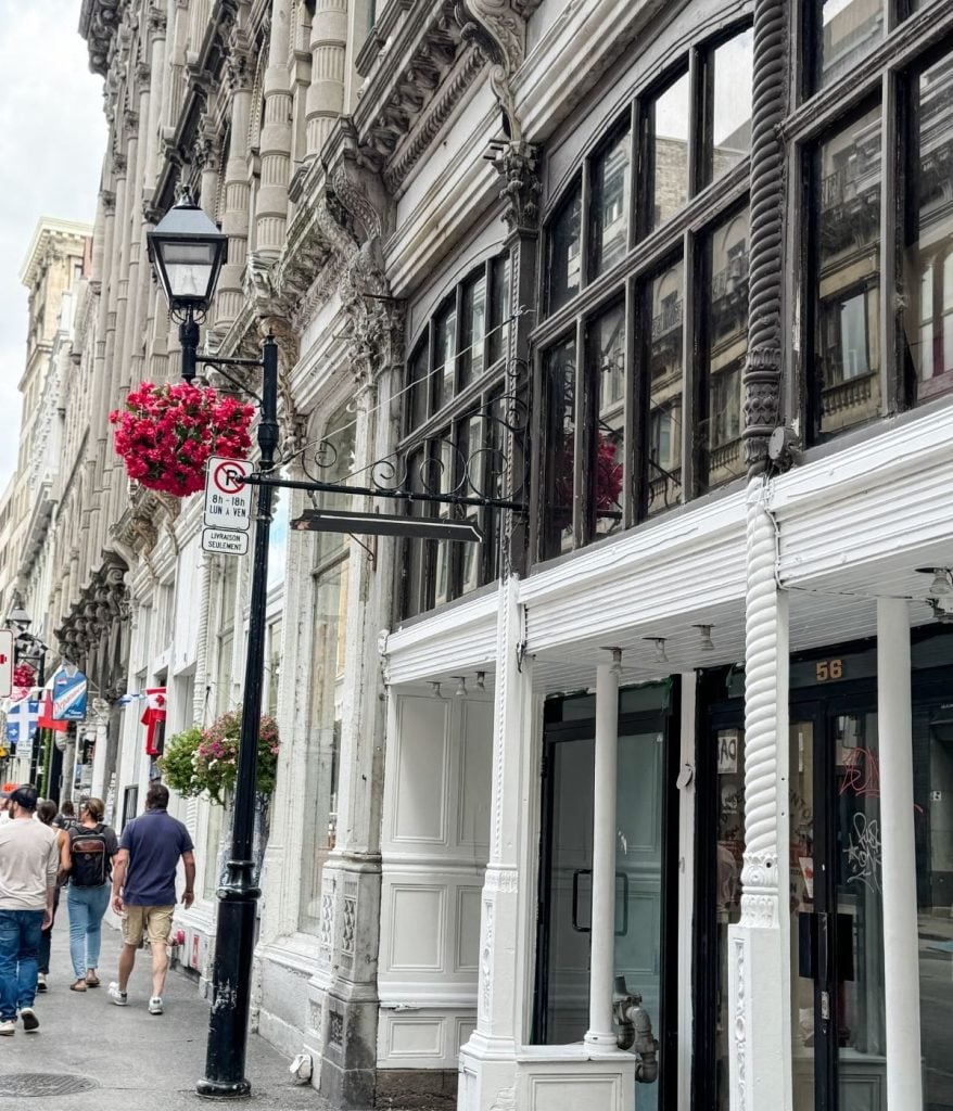 A city street with ornate historic buildings, hanging flower baskets, a lamp post, and pedestrians—one sporting a stylish men's crochet cardigan—walking on the sidewalk.