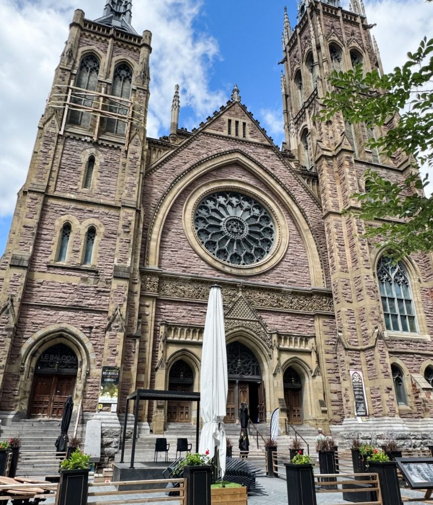 Gothic-style church with two tall towers, a large circular stained glass window, and arched entrance, with outdoor seating where a men's crochet cardigan adds charm among the closed umbrellas in front.