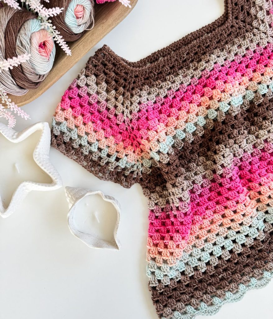A crocheted Old Town Tee featuring brown, pink, red, and light blue yarn is displayed on a white surface next to a tray of yarn balls and two star-shaped items. This piece highlights the timeless Granny Stitch technique.