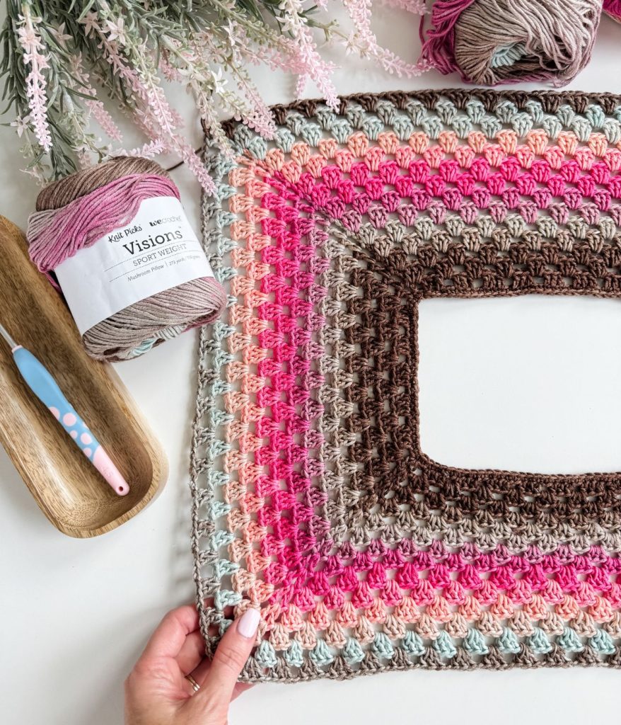 A hand holds a partially finished crochet granny square in pink, brown, gray, and cream tones; yarn, a crochet hook, and artificial flowers are nearby—perfect inspiration for your next Granny Stitch Top or Old Town Tee project.