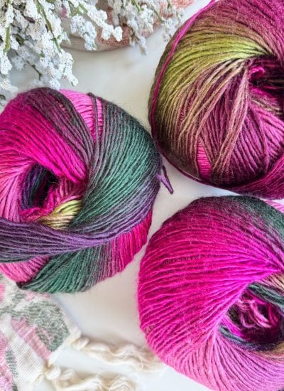 Three skeins of multicolored yarn in shades of pink, purple, and green are arranged on a flat surface with decorative white foliage nearby—perfect for starting your next Old Town Sweater project.