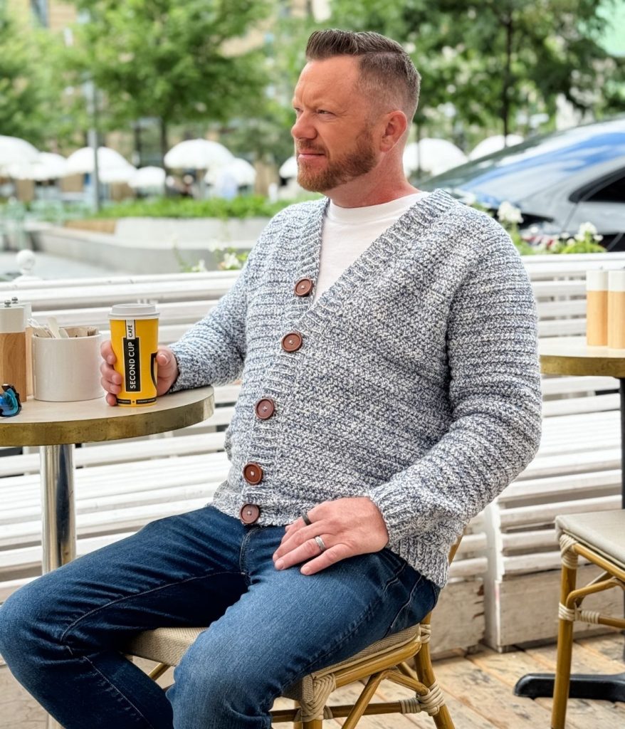 A man in a men's crochet cardigan and jeans sits at an outdoor café table, holding a can of lemon craft soda, with trees and parked cars in the background.