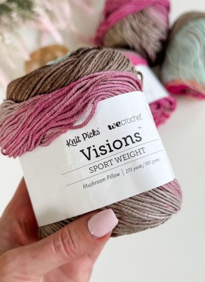 A hand holds a skein of 'Visions' sport weight yarn in the color 'Mushroom Pillow' by Knit Picks and WeCrochet—perfect for your next Old Town Tee or Granny Stitch top pattern; more skeins appear in the background.