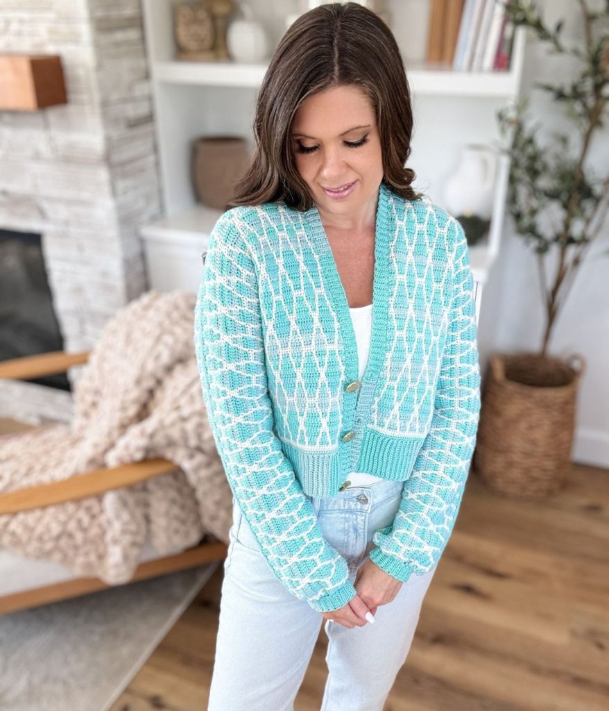Woman with long brown hair wearing a light blue Seaglass mosaic cardigan and white top, standing indoors on wooden flooring near a chair and shelves.
