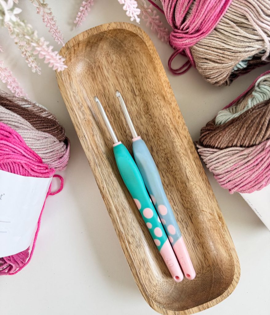 Two crochet hooks with colorful handles lie in a wooden tray, surrounded by skeins of pink, brown, and beige yarn—perfect for starting your next Granny Stitch Top or following a free pattern like the Old Town Tee.