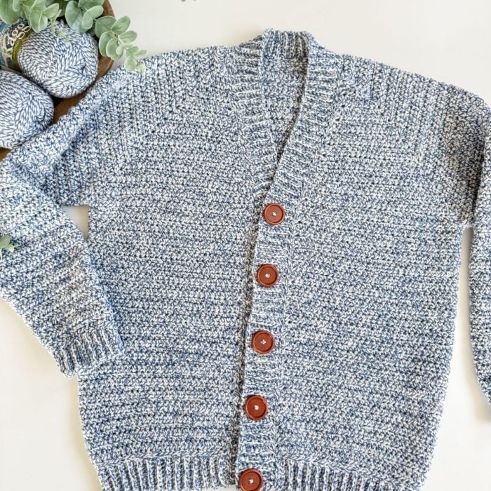 A blue and white men's crochet cardigan with brown buttons is laid flat on a white surface next to a basket containing yarn and green foliage.