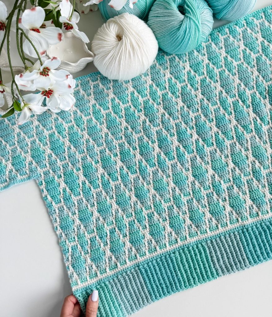 A hand holds a knitted turquoise and white patterned fabric, reminiscent of a Mosaic Cardigan, with matching yarn balls and white flowers arranged nearby on a flat surface.