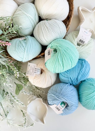 Balls of pastel-colored yarn in white, mint, and blue shades—perfect for crafting a Mosaic Cardigan—are arranged on a table with decorative greenery and star-shaped candles.