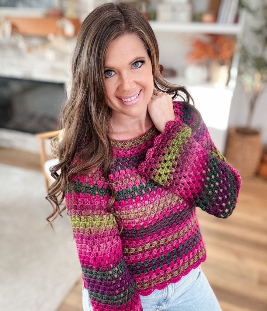 A woman with long brown hair wears the vibrant Old Town Sweater—crocheted in shades of pink, green, and purple—standing indoors and smiling at the camera.