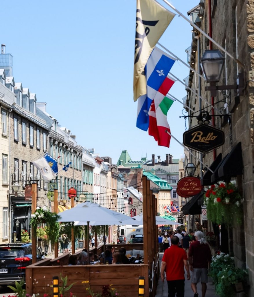 People walk along a historic street lined with shops, restaurants, and flags, including the Quebec flag, on a sunny day. In the foreground, a man wearing a stylish men's crochet cardigan enjoys outdoor patio seating.