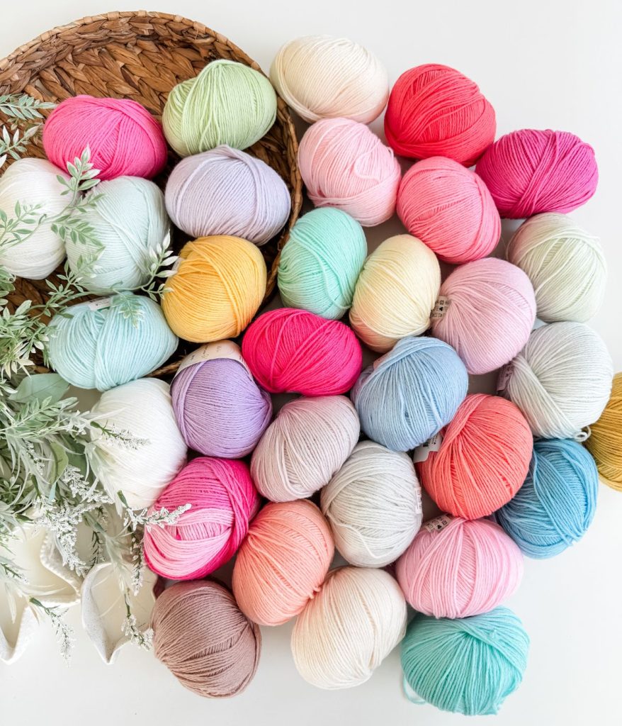 A variety of colorful yarn balls are arranged in a neat cluster on a white surface, perfect for your next Mosaic Cardigan project, with greenery and a woven mat partially visible on the left side.