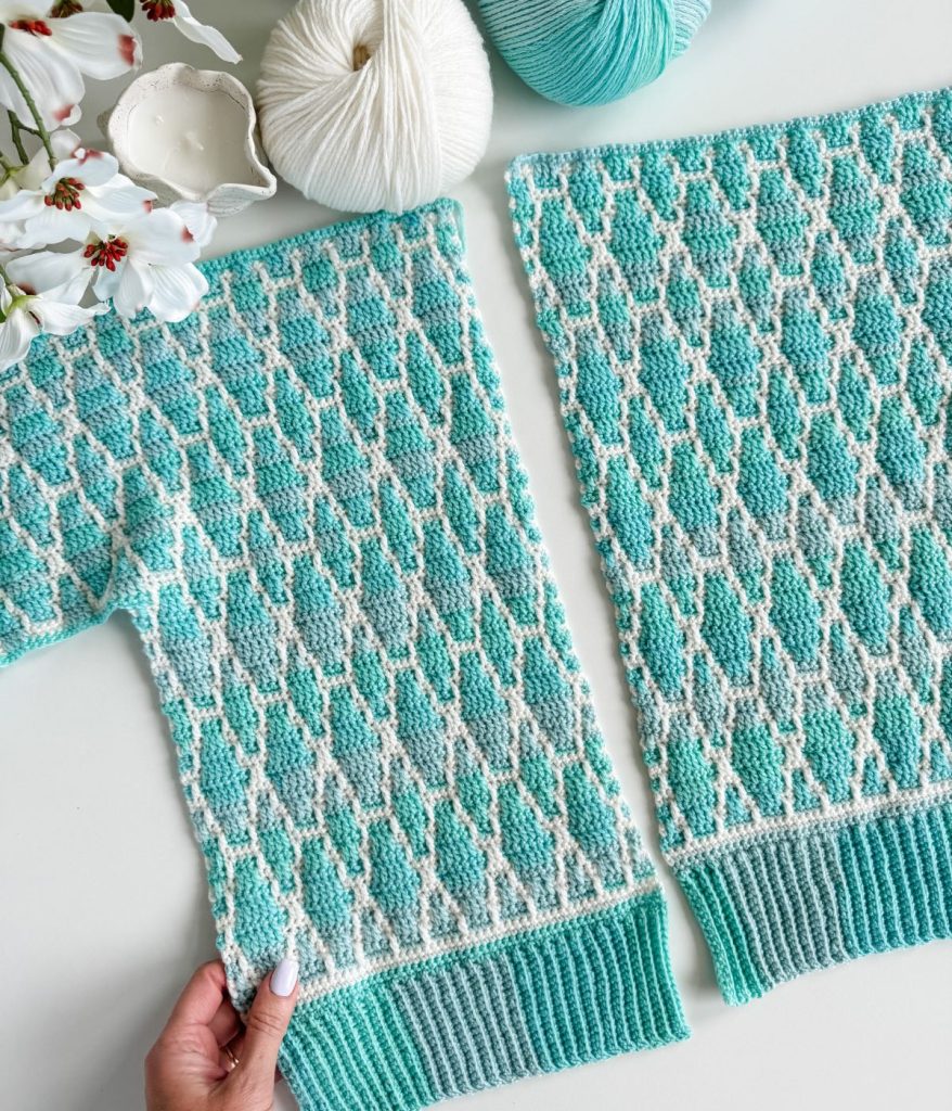 Two knitted panels with a turquoise and white geometric pattern, inspired by the Seaglass Mosaic Cardigan, are displayed on a white surface beside yarn balls and white flowers. A hand holds one panel at the bottom edge.