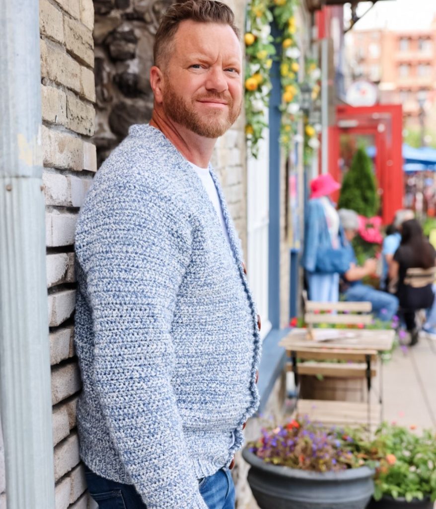 A man with a beard and light blue men's crochet cardigan stands against a brick wall on a lively street with flowers and people in the background.
