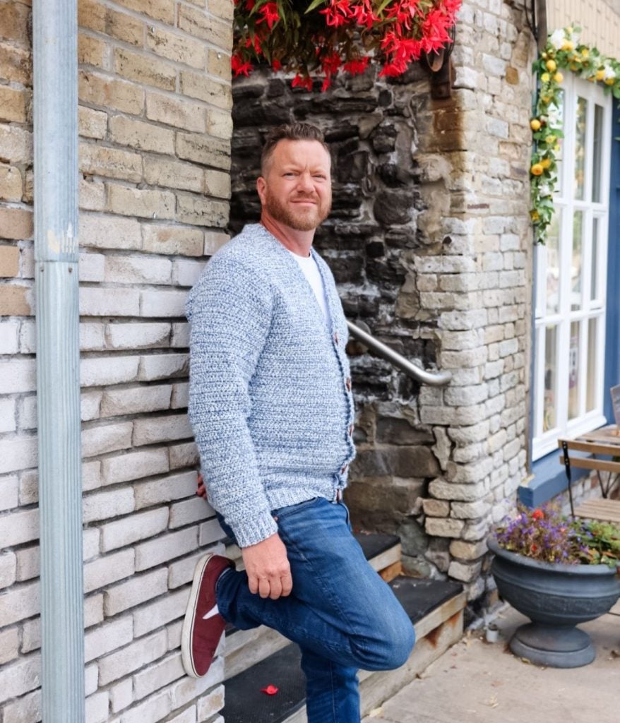 A man wearing a men's crochet cardigan, jeans, and red sneakers leans against a brick wall outdoors, with flowers and a window beside him.