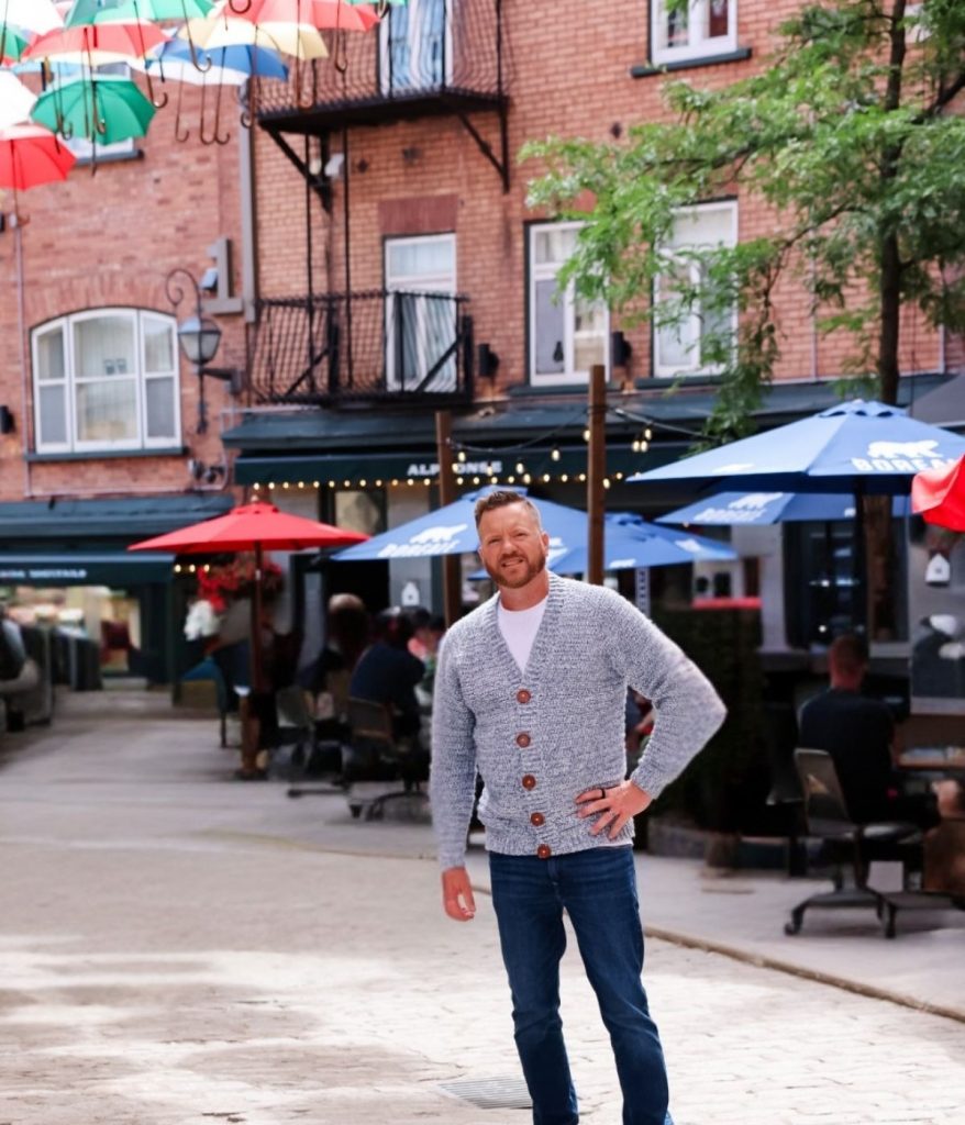 A man in a men's crochet cardigan stands on a cobblestone street lined with outdoor seating, colorful umbrellas, and brick buildings in the background.