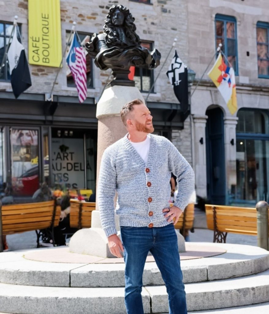 A man in a stylish men's crochet cardigan and jeans stands in front of a statue in a city square, with flags and a café visible in the background.