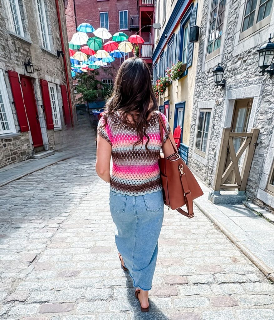 A woman with long brown hair, wearing a granny stitch top and denim skirt, walks down a cobblestone street with colorful umbrellas suspended overhead.