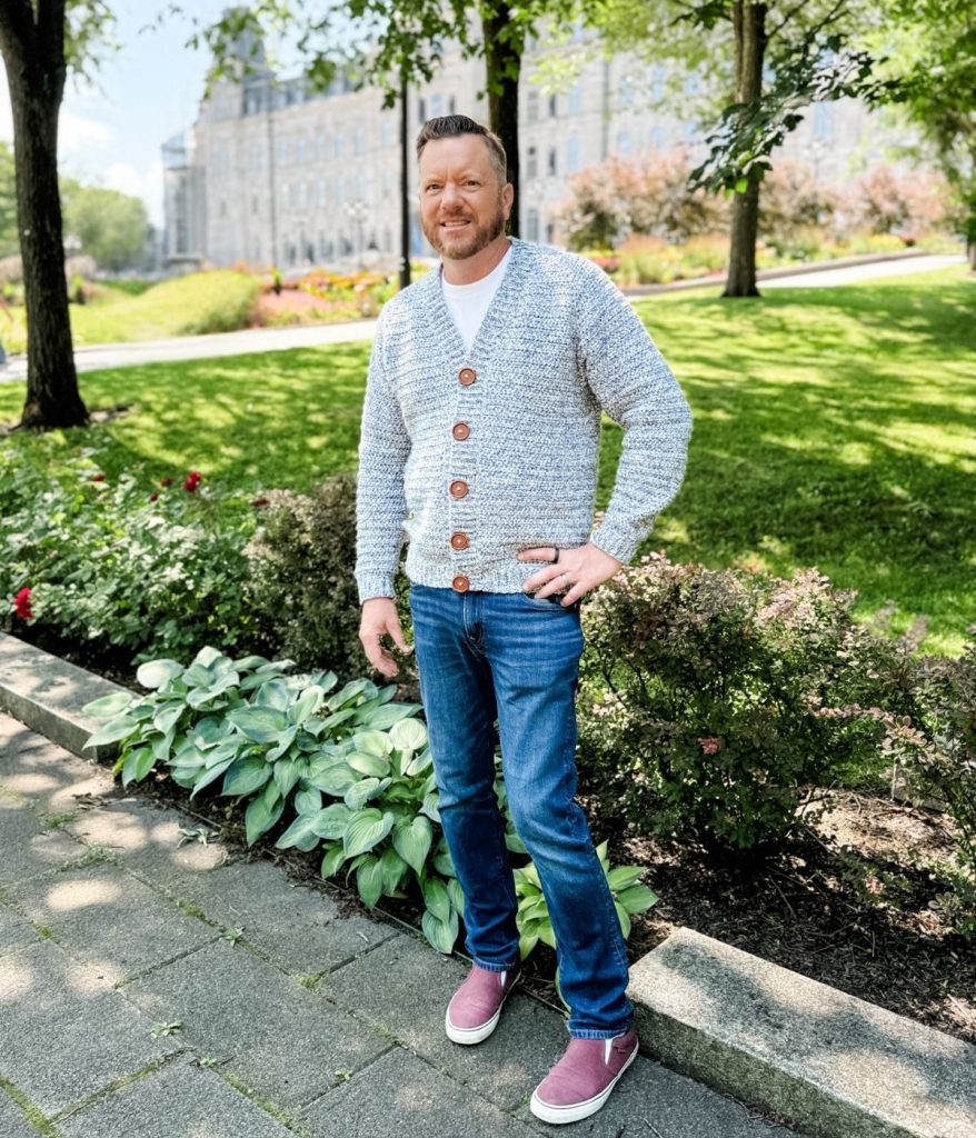 A man stands on a stone path in a park, wearing a stylish men's crochet cardigan, blue jeans, and maroon slip-on shoes, with lush greenery and a large building in the background.