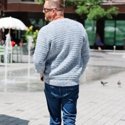 A man wearing a men's crochet cardigan and blue jeans walks away on a paved area near fountains and trees on a sunny day.