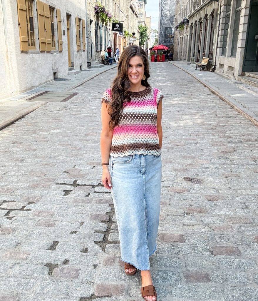 A woman wearing a striped Granny Stitch Top and light blue jeans stands and smiles on a cobblestone street lined with historic buildings.