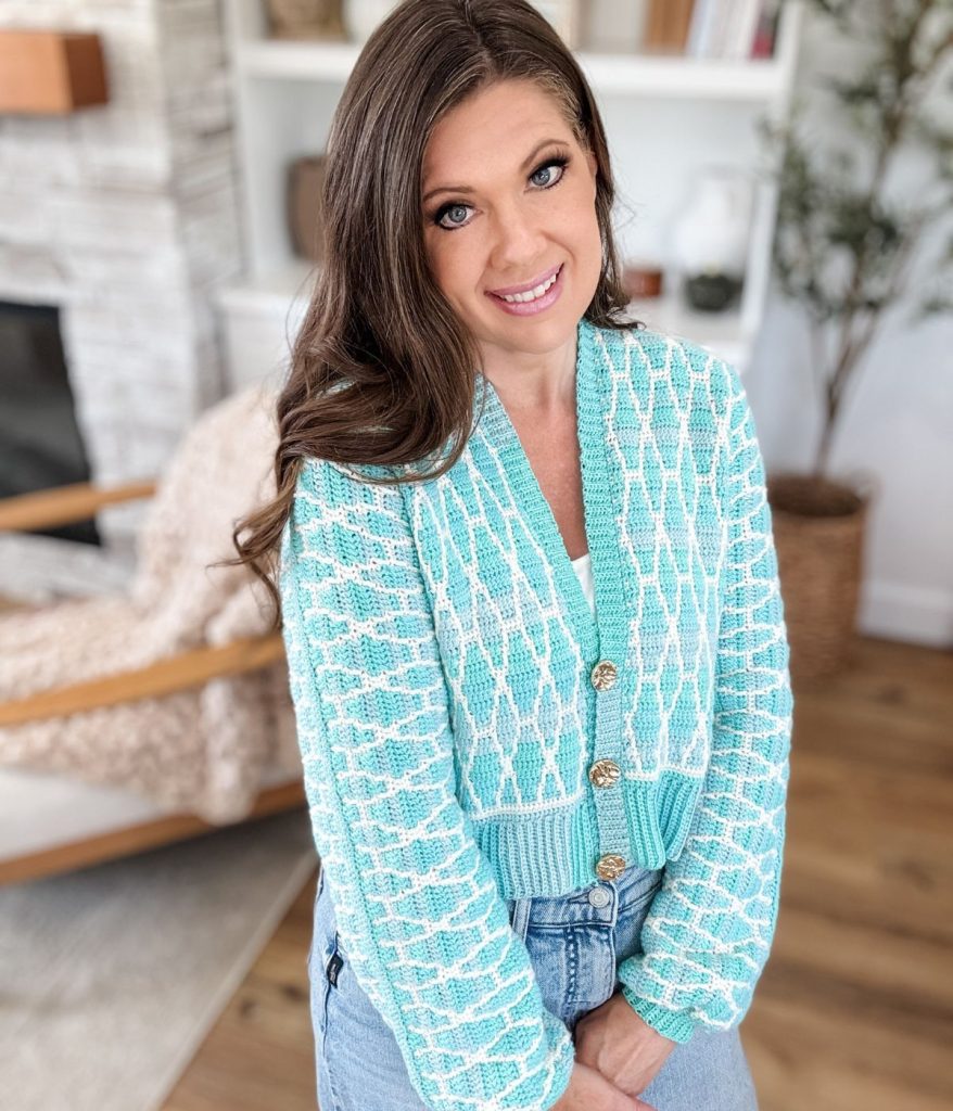 Woman with long brown hair wearing a Seaglass Mosaic Cardigan and jeans, standing indoors in a bright living room with wooden floors and shelves in the background.