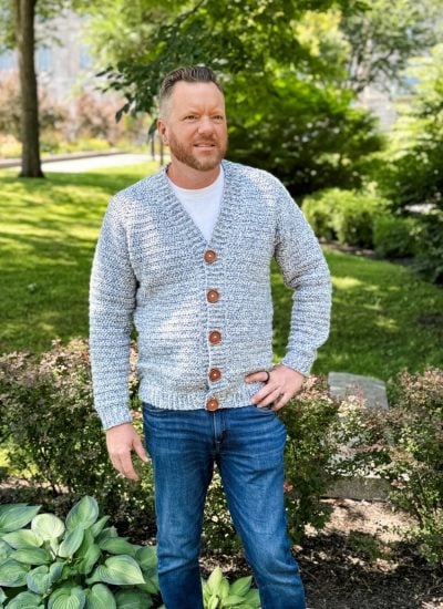 A man stands outdoors in a park, wearing a light gray men's crochet cardigan with brown buttons, a white t-shirt, and blue jeans. Green plants and trees are visible in the background.