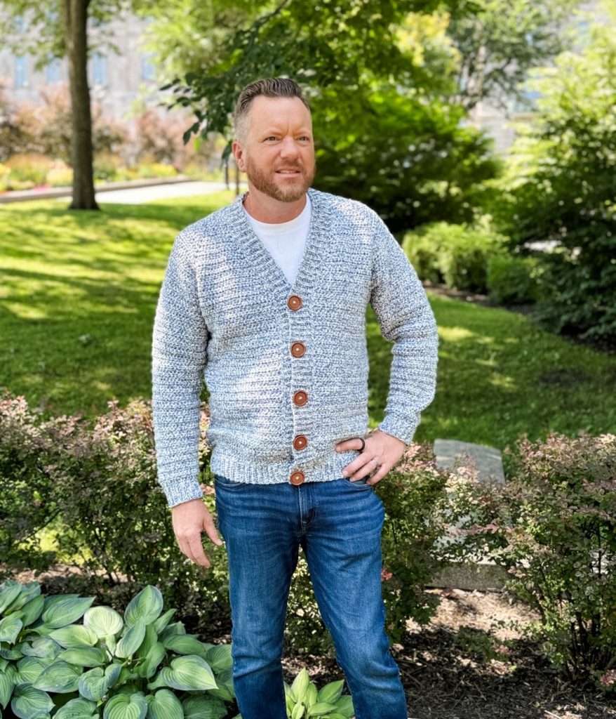 A man stands outdoors in a park, wearing a light gray men's crochet cardigan with brown buttons, a white t-shirt, and blue jeans. Green plants and trees are visible in the background.