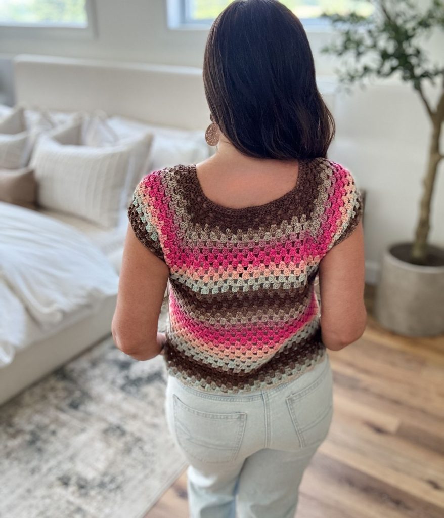 A woman stands indoors facing away, wearing a crocheted granny stitch top—the Old Town Tee—with brown, pink, and cream stripes and light-colored jeans. A bed and potted plant are visible in the background.