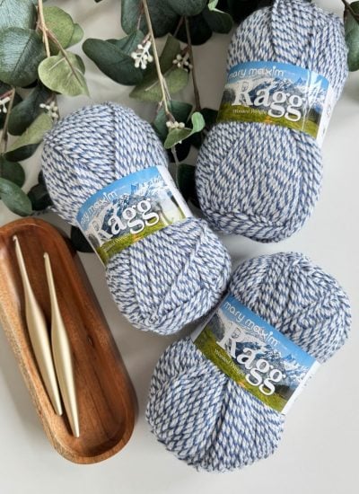 Three skeins of blue and white marled yarn, two crochet hooks in a wooden tray, and green foliage on a white surface—perfect supplies for crafting a stylish men's crochet cardigan.