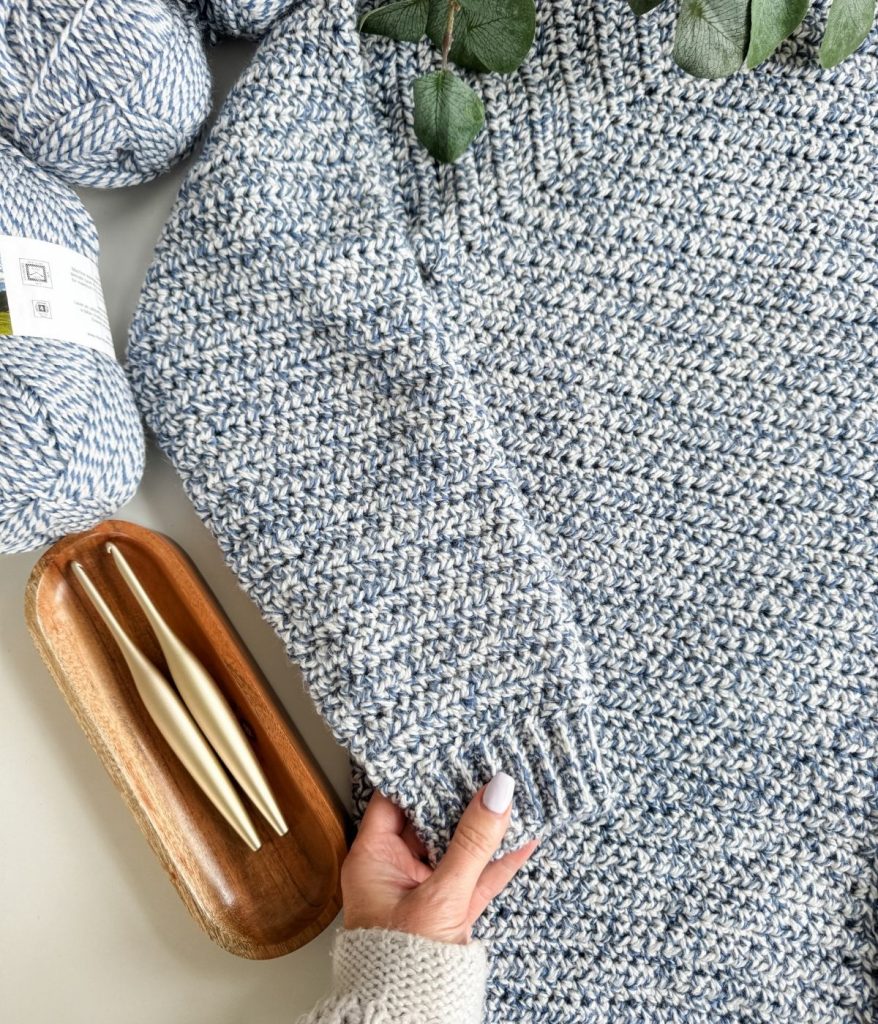 A hand holds the sleeve of a blue and white Men's Crochet Cardigan, with yarn balls, two crochet hooks in a wooden tray, and green leaves nearby.