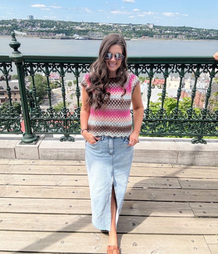 Woman wearing sunglasses, a stylish Granny Stitch Top with stripes, and a long denim skirt stands on a wooden deck with a river and cityscape in the background.