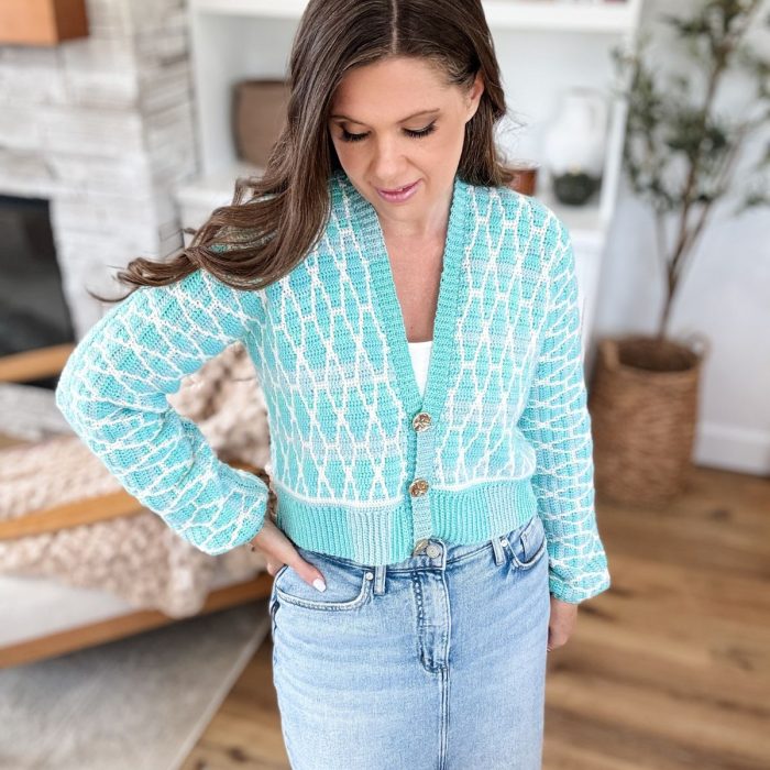 Woman standing indoors wearing a Seaglass Mosaic Cardigan over a white top and light blue jeans, looking down with one hand in her pocket.