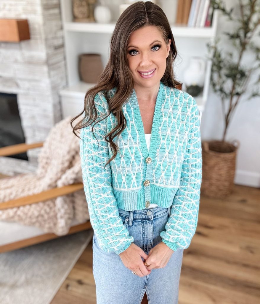 Woman with long brown hair wearing a light blue seaglass-patterned cardigan and denim skirt stands indoors, smiling at the camera. Shelves and a chair are visible in the background.