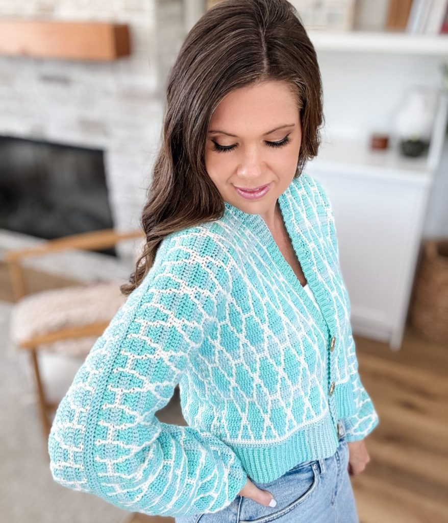 Woman with long brown hair wears a Seaglass Cardigan in light blue and white patterns with high-waisted jeans, standing indoors and looking down.