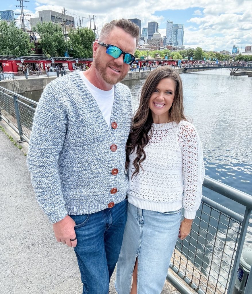 A man and a woman stand side by side by a waterfront railing, both smiling at the camera. The man is wearing a stylish men's crochet cardigan, while buildings and people can be seen in the background.
