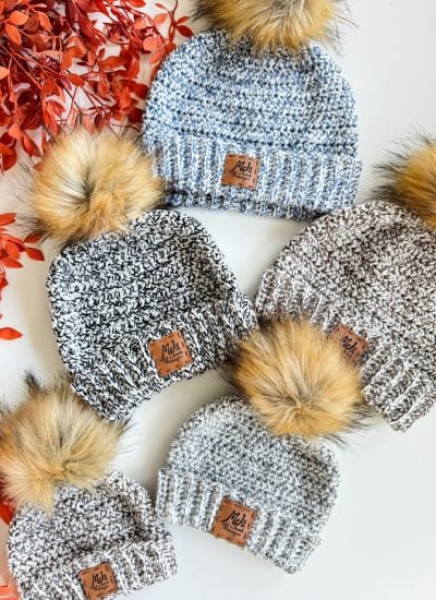 Five knit beanies with faux fur pom-poms and brown labels are arranged on a white surface next to red-orange artificial leaves, offering inspiration for your next easiest crochet beanie or free crochet pattern project.