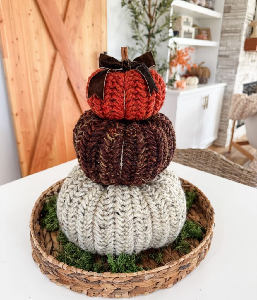 Three knitted pumpkins, including a charming Herringbone Pumpkin in orange, brown, and cream, are stacked on a wicker tray with green moss accents, set on a white table in a cozy, decorated room.