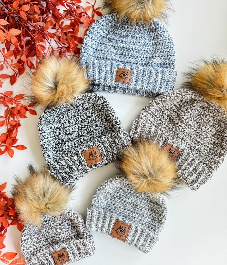 Five knit beanies with faux fur pom-poms and brown labels are arranged on a white surface next to red-orange artificial leaves, offering inspiration for your next easiest crochet beanie or free crochet pattern project.
