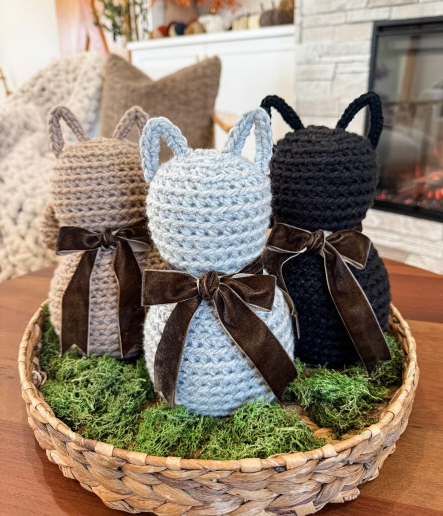 Three Crochet Cat figures in brown, gray, and black, each with a brown bow, are displayed in a wicker basket lined with moss on a wooden table.