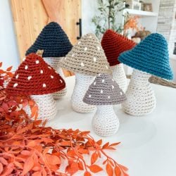 Six Crochet Mushroom figures with white stems and red, blue, beige, or gray spotted caps are arranged on a tabletop next to orange foliage in a modern living space.