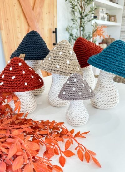 Six Crochet Mushroom figures with white stems and red, blue, beige, or gray spotted caps are arranged on a tabletop next to orange foliage in a modern living space.