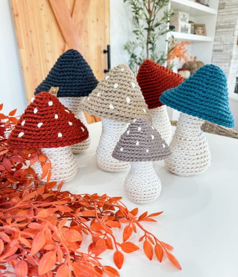 Six Crochet Mushroom figures with white stems and red, blue, beige, or gray spotted caps are arranged on a tabletop next to orange foliage in a modern living space.