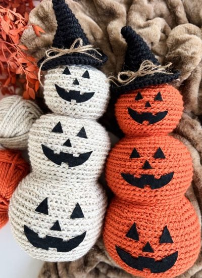 Two crocheted stacked pumpkins—one white, one orange—feature classic Crochet Jack-O-Lantern faces in black felt and wear black witch hats, all displayed on a brown textured fabric.