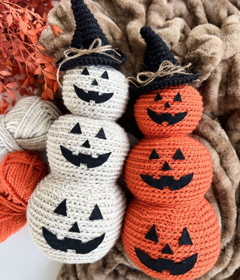 Two crocheted stacked pumpkins—one white, one orange—feature classic Crochet Jack-O-Lantern faces in black felt and wear black witch hats, all displayed on a brown textured fabric.
