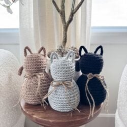 Three adorable Crochet Cat figures in beige, gray, and black with twine bows sit on a small round wooden table in front of a potted plant.