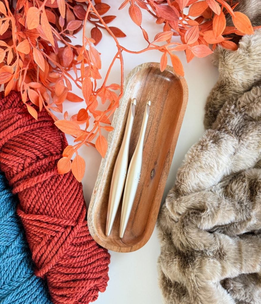 Two crochet hooks in a wooden tray, surrounded by red yarn, blue yarn, faux fur fabric, orange artificial leaves, and a charming crochet mushroom for a cozy crafting vibe.